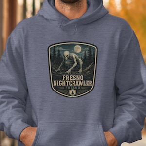 May include: Blue hoodie featuring a graphic of the Fresno Nightcrawler, a cryptid, in a forest scene under a full moon. The design includes the text "FRESNO NIGHTCRAWLER" and "FRESNO". The hoodie is a solid blue color.