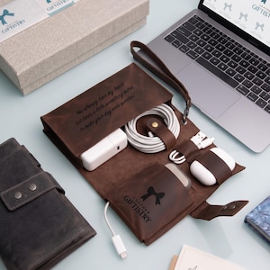 May include: A dark brown leather tech organizer roll featuring a wrist strap, internal pockets, and elastic loops holding white charging cables, a power adapter, and wireless earbuds. The leather is engraved with the text: You already have my heart, but here's a little something extra to make your day even sweeter!
