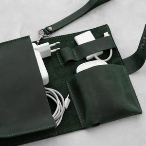 May include: Dark green leather tech organizer with multiple compartments. The open organizer displays a white charging brick, earbuds, and a charging cable. A wrist strap and snap closure are visible.