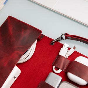 May include: A red leather travel case with a strap, designed to hold and organize various electronic accessories, including charging cables, a USB drive, and a pair of wireless earbuds.