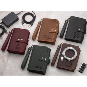 May include: Five rectangular leather tech organizers in burgundy, brown, black, dark green, and dark brown. Each features a wrist strap, a snap-button closure, and personalized engravings. One organizer holds a white coiled charging cable.