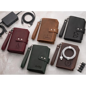 May include: Five rectangular leather tech organizers in burgundy, brown, black, dark green, and dark brown. Each features a wrist strap, a snap-button closure, and personalized engravings. One organizer holds a white coiled charging cable.