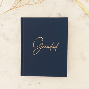 Puede incluir: Un diario azul marino con la palabra "Grandad" en escritura dorada. El libro cerrado está centrado. El fondo es una superficie de mármol claro con una ramita de flores secas y dos bolígrafos.