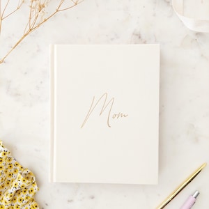 May include: A white journal with the word "Mom" written in gold script on the front cover. The journal is on a white marble surface with a sprig of dried flowers and a pink and gold pen.