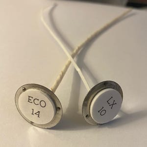 May include: Two candle wicks with circular bases. One base is labeled "ECO 14" and the other "LX 10". The bases are white with a silver metal rim. The wicks are white and beige.