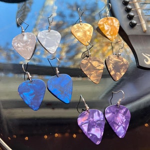 May include: Eight pairs of guitar pick earrings in various colors, including white, gold, brown, blue, and purple. The earrings are displayed on a black surface with a guitar in the background.