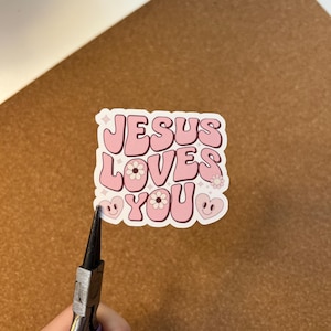 May include: A pink sticker with the words "JESUS LOVES YOU" in a retro font. The sticker features floral and heart designs. The sticker is being held by a pair of pliers.