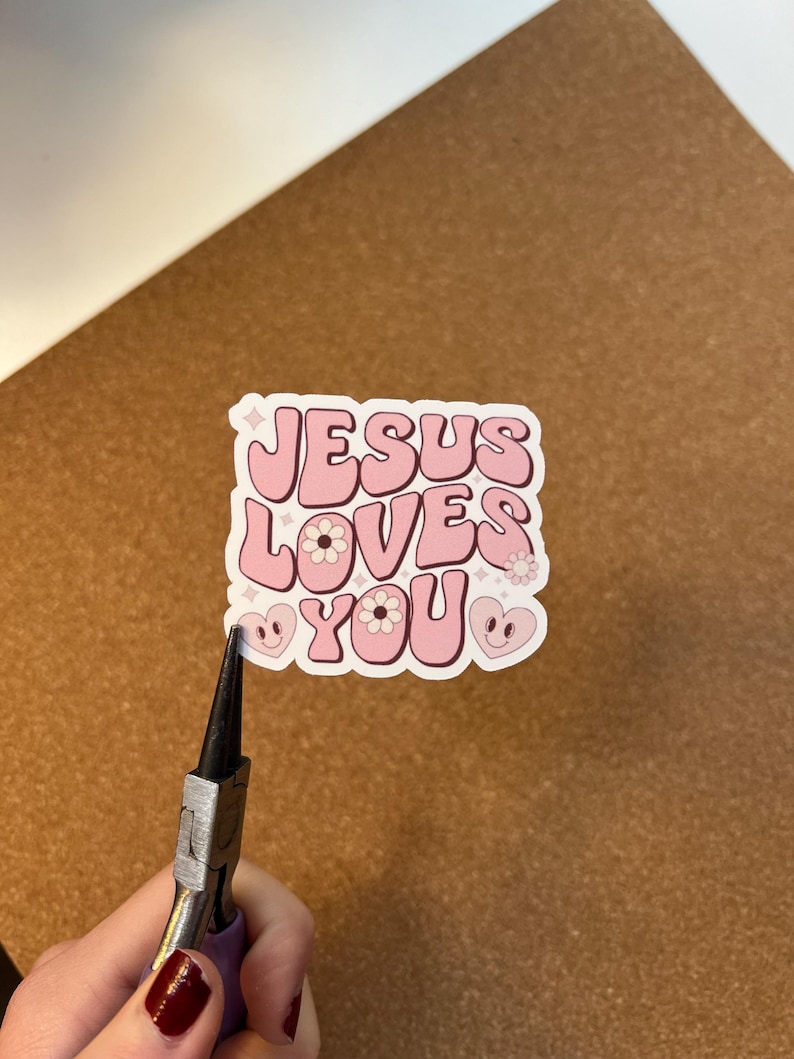 May include: A pink sticker with the words "JESUS LOVES YOU" in a retro font. The sticker features floral and heart designs. The sticker is being held by a pair of pliers.
