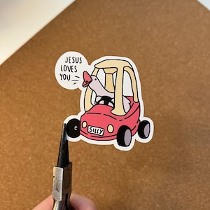 May include: A sticker showing a cartoon goose in a red toy car with the word "SILLY" on the front. A speech bubble says "JESUS LOVES YOU." The sticker is on a brown surface.