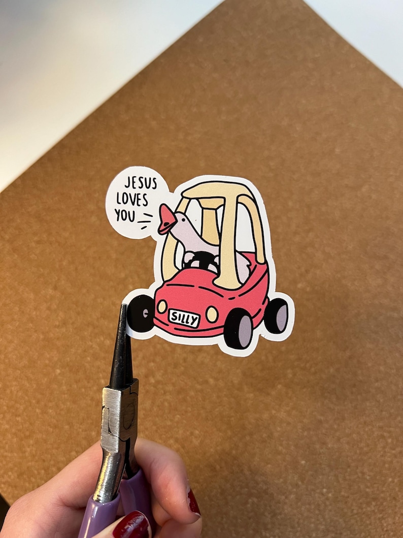 May include: A sticker showing a cartoon goose in a red toy car with the word "SILLY" on the front. A speech bubble says "JESUS LOVES YOU." The sticker is on a brown surface.