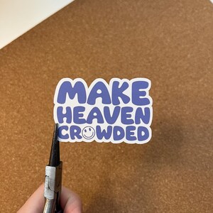 May include: A sticker with the text "MAKE HEAVEN CROWDED" in blue letters with a white outline. The word "HEAVEN" has a smiling face in place of the letter "O". The sticker is being held by pliers.