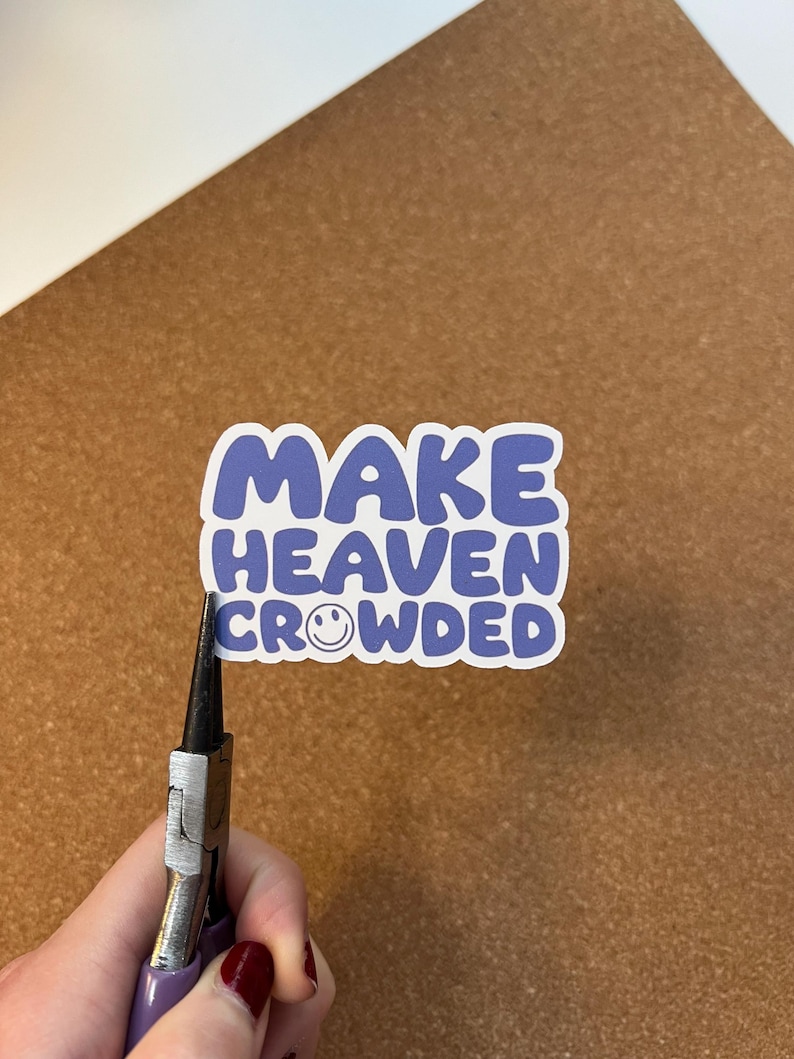 May include: A sticker with the text "MAKE HEAVEN CROWDED" in blue letters with a white outline. The word "HEAVEN" has a smiling face in place of the letter "O". The sticker is being held by pliers.