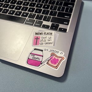 May include: A laptop with stickers. One sticker reads "NEWS FLASH" with a cross and the text "God is still on the throne!". Another sticker shows a jar labeled "THE GOSPEL" with a knife spreading pink jam on toast. The text "GO SPREAD IT" is also visible.