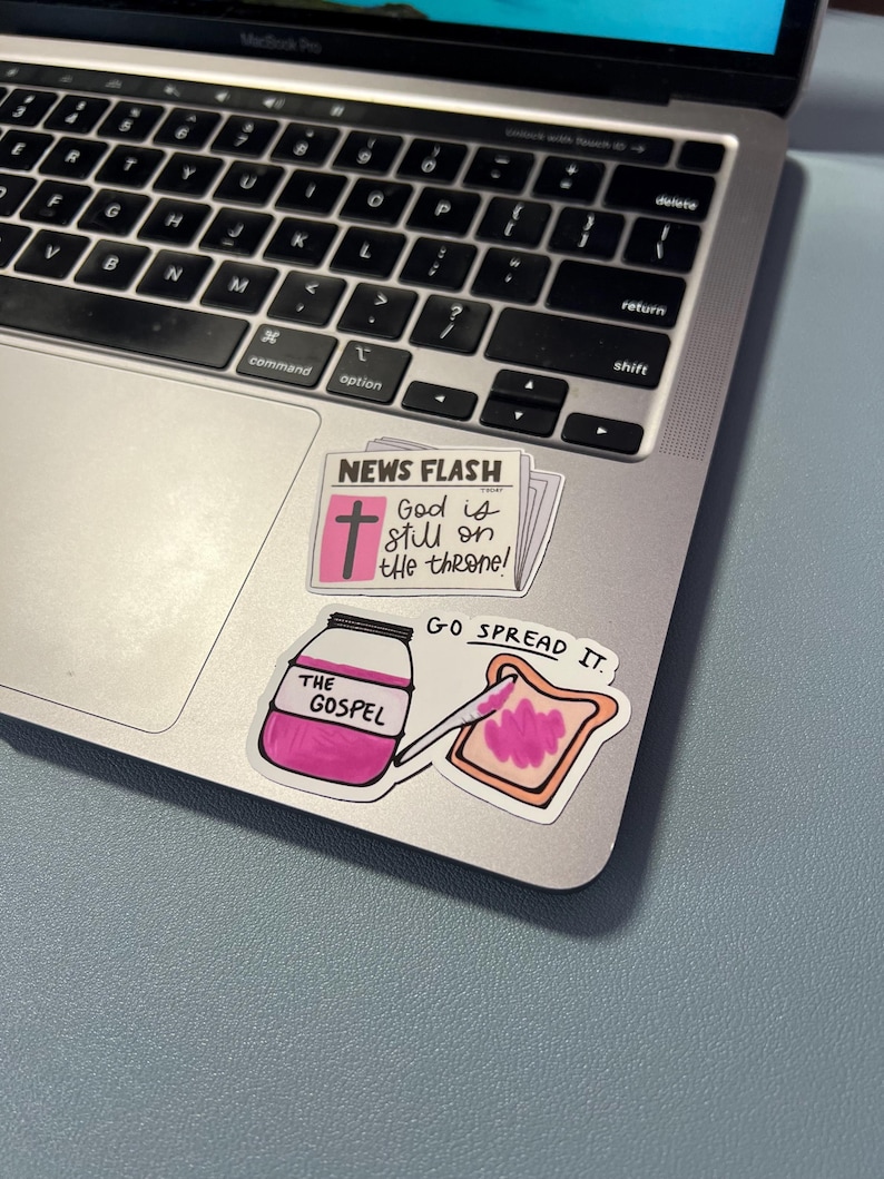May include: A laptop with stickers. One sticker reads "NEWS FLASH" with a cross and the text "God is still on the throne!". Another sticker shows a jar labeled "THE GOSPEL" with a knife spreading pink jam on toast. The text "GO SPREAD IT" is also visible.