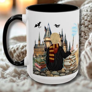 Könnte beinhalten: Weißer Keramikbecher mit schwarzem Innenraum und Henkel. Der Becher zeigt eine farbenfrohe Illustration einer Person in schwarzem Umhang und Schal, die einen Zauberstab hält, mit einer Burg und Büchern im Hintergrund. Der Becher steht auf einer weichen, strukturierten Oberfläche.