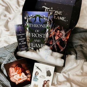 May include: A black box with a white interior, containing a book titled "A Throne of Frost and Flame" with a blue cover and a silver design. The box also contains a bookmark, a sticker sheet, and a print of a couple embracing.
