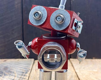 May include: A handmade robot sculpture crafted from wood and metal components. The robot features a red painted wooden head and body, with metal hardware used for eyes, arms, and legs. The robot's arms are made of springs and metal fasteners. The robot stands on a wooden surface.