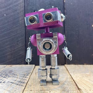 May include: A small, purple robot made of metal and wood. The robot has two round eyes, a round head, and a rectangular body. The robot is standing on a wooden surface.