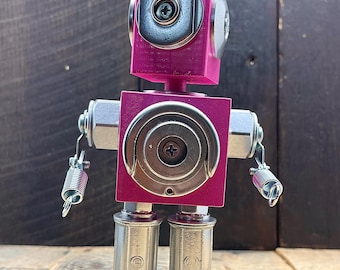 May include: A handmade robot sculpture with a magenta painted wooden head and body. The robot's head and body are square-shaped, with metal hardware accents, including bolts, washers, and springs for arms. The legs are made of cylindrical metal pieces.