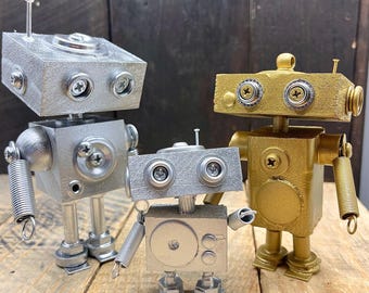 May include: Three robot figurines made of metal and painted silver and gold. The robots are standing on a wooden surface.
