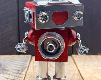 May include: A handcrafted robot sculpture made from repurposed metal and wood. The robot features a red head and body, with silver metal accents for the face, arms, and legs. The robot's arms are made of wire and the legs are made of metal pipes.