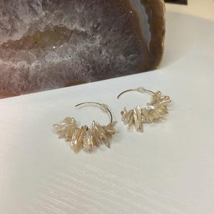 May include: A pair of hoop earrings with a cluster of irregular, light-colored, elongated beads. The earrings are set against a white surface, with a large, textured, purple-grey stone in the background. The earrings are a jewelry item.