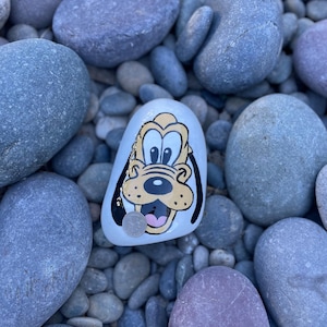 May include: A painted rock with a cartoon image of a dog with brown fur, a black nose, and white eyes. The dog is smiling and has a pink tongue sticking out. The rock is surrounded by other rocks.