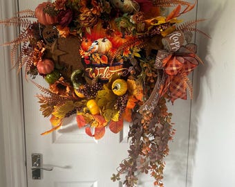 May include: A fall-themed wreath with pumpkins, sunflowers, and autumn leaves. The wreath includes a scarecrow hat and a ribbon with the word "Happy". The wreath is a mix of orange, brown, and yellow colors.