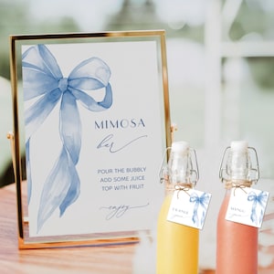 May include: A gold-framed sign with a blue watercolor bow and the words "Mimosa Bar." Two glass bottles with swing-top closures and tags that read "Orange Juice" and "Mango Juice" are in front of the sign.