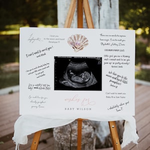 Puede incluir: Un letrero blanco sobre un caballete de madera, con el texto "wishes for BABY WILSON" en escritura rosa. El letrero incluye una imagen de ultrasonido, una ilustración de concha y mensajes manuscritos.