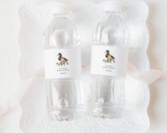 May include: Two clear plastic water bottles with white labels featuring a watercolor duck illustration. The labels read "LAUREN'S BABY SHOWER" and a date. The bottles are on a white decorative tray with a ribbon.