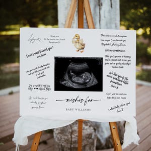 Puede incluir: Un letrero blanco sobre un caballete con deseos para el baby shower. El letrero incluye una imagen de ultrasonido, un patito de acuarela y mensajes escritos a mano. El texto dice "wishes for BABY WILLIAMS."