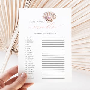 May include: A baby shower game card with the words "Baby Word Scramble" in pink script. The card features a seashell graphic and a list of scrambled words to unscramble. The card is held by a hand.