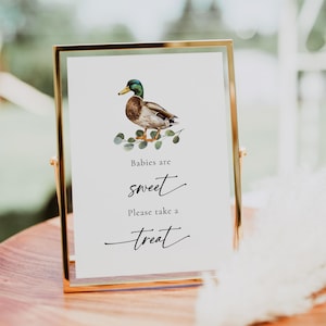 May include: A gold-framed sign with a watercolor illustration of a duck and eucalyptus leaves. The sign reads "Babies are sweet. Please take a treat."