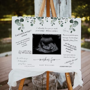 Puede incluir: Un letrero blanco sobre un caballete con detalles de hojas de eucalipto y el texto "wishes for BABY WILLIAMS". El letrero incluye una imagen de ultrasonido y mensajes escritos a mano como "I can't wait to meet you!" y "I already love you so much sweet baby!"