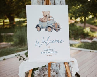 May include: Welcome to Avery's Baby Shower sign with a watercolor illustration of a teddy bear driving a blue car. The sign is on a wooden easel and has a white background with blue and brown text.