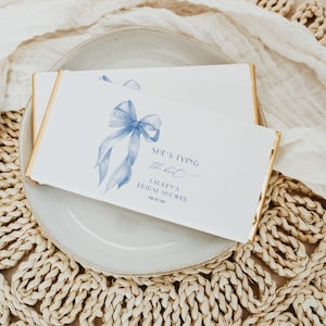 Könnte beinhalten: Eine weiße Karte mit einer hellblauen Aquarellschleife und dem Text "She's tying the knot. Lauren's Bridal Shower 08.01.XX". Die Karte hat goldene Ränder und liegt auf einem weißen Teller.