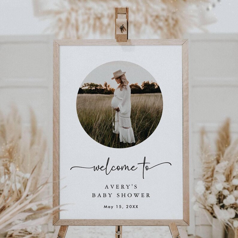 May include: Welcome to Avery's Baby Shower sign with a photo of a pregnant woman in a white dress and hat standing in a field. The sign is on a wooden easel and has a white background with black text. The date of the shower is May 15, 20XX.