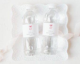 May include: Two clear plastic water bottles with white labels and pink bow illustrations. The labels read "Bridal Shower" with a date. The bottles sit on a white, decorative tray with a ribbon.