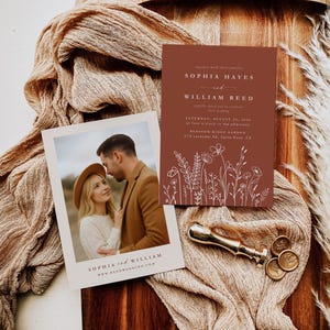 May include: A wedding invitation suite with a photo card and a terracotta-coloured invitation featuring white floral illustrations. The photo card shows a couple, while the invitation includes event details. A gold wax seal and decorative fabric complete the scene.