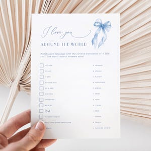 Puede incluir: Una tarjeta blanca con el texto "I love you AROUND THE WORLD" en escritura azul. La tarjeta presenta un juego de emparejamiento con traducciones de "I love you" en varios idiomas. Un lazo azul de acuarela está en la esquina superior derecha.