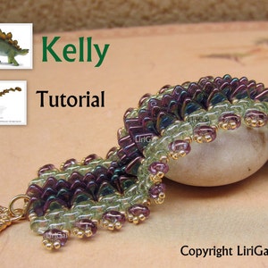 Puede incluir: Un brazalete de cuentas verdes y moradas con detalles dorados. El brazalete está hecho con pequeñas cuentas de semillas y presenta un diseño único y escamoso. El brazalete se muestra sobre una superficie marrón claro con una pequeña piedra.