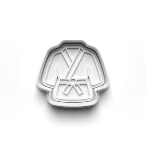 May include: A white plastic cookie cutter shaped like a karate gi. The cutter features a detailed outline of the jacket, belt, and lapels. The design is clean and simple, ideal for making themed biscuits.