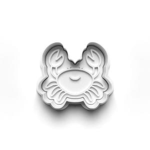 May include: A white plastic crab-shaped cookie cutter. The cutter has a smiling crab face, two claws, and a scalloped edge. The design is simple and clean, ideal for baking biscuits.