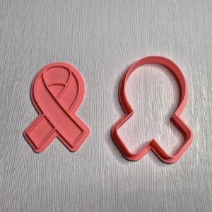 May include: Two pink ribbon-shaped cookie cutters. One is a solid ribbon design, and the other is the outline of the ribbon. The cutters are made of plastic and are on a light grey textured surface.