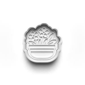 May include: A white plastic cookie cutter shaped like a flower pot with two flowers and leaves. The cutter has a scalloped edge, designed for baking. The design is simple and clean, ideal for creating floral-themed biscuits.
