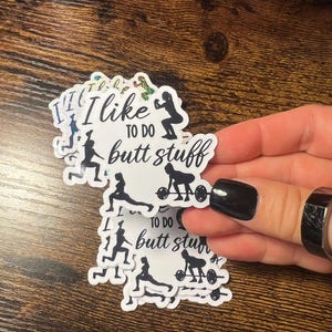May include: A collection of white stickers with the phrase "I like to do butt stuff" in black script. The stickers feature silhouettes of people exercising with weights and doing squats. The stickers are held by a hand with a black painted nail.