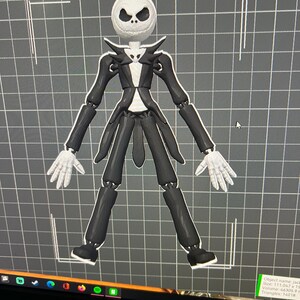May include: A black and white articulated figure of Jack Skellington from the movie The Nightmare Before Christmas. The figure is wearing a black suit with a white shirt and a black bow tie. The figure is standing on a grid background.