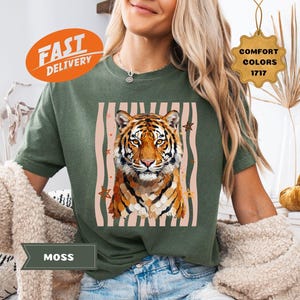 May include: A moss green t-shirt featuring a tiger graphic with a striped background. The tiger has orange and black stripes. The shirt has the words "Comfort Colors 1717" on a tag. The words "Fast Delivery" are on an orange oval.