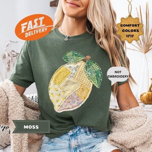 May include: A moss green t-shirt featuring a patchwork lemon design. The lemon is made of various patterned fabrics in yellow, green, and floral prints. The shirt has the words "Fast Delivery" on it.